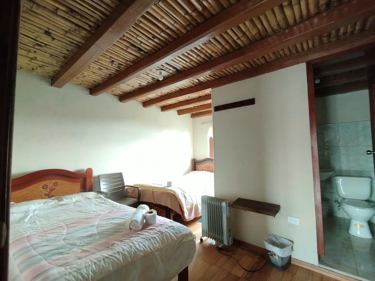 Quarto com teto de madeira, cama com cabeceira floral, radiador e vaso sanitário visível por uma porta de vidro.