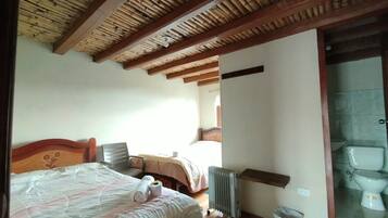 Quarto com teto de madeira, cama com cabeceira floral, radiador e vaso sanitário visível por uma porta de vidro.