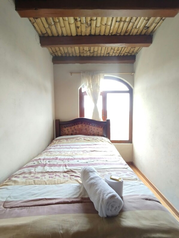 Una cama individual con una toalla enrollada, una ventana con cortinas y un techo de bambú.