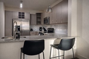 A modern kitchen with stainless steel appliances, a granite countertop, and two black bar stools. - Vibrant DT Condo NR Farmers Market (Dallas)