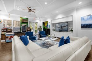 Uma sala de estar moderna com um sofá de couro branco, poltronas azuis e uma mesa de centro. Há uma televisão na parede e um ventilador de teto.