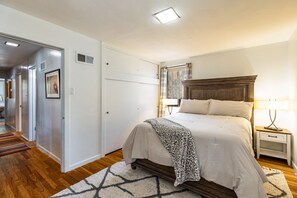 Quarto com cama, mesas de cabeceira, luminária, janela com cortinas, porta e corredor que dá acesso a outros ambientes.