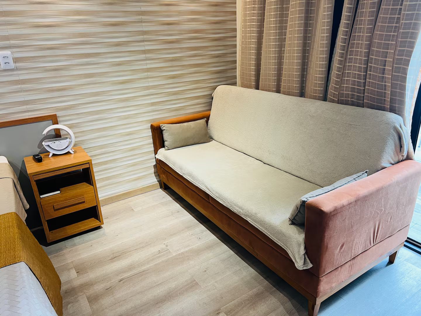Una habitación con piso de madera, un sofá, una mesita de noche con lámpara y una ventana con cortinas.