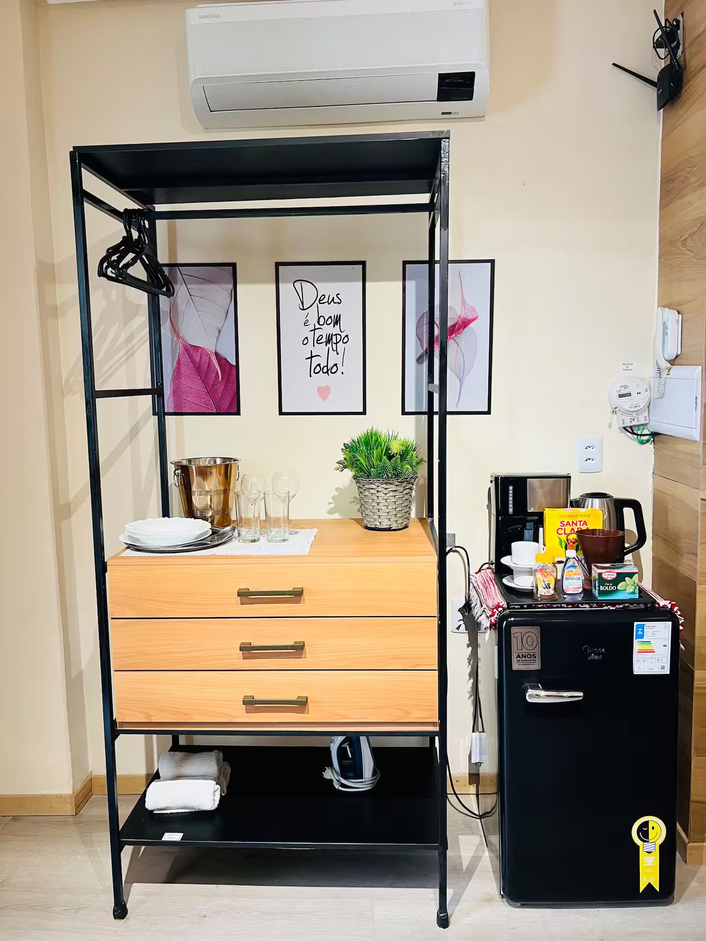 Quarto compacto com cama, uma mesinha com uma planta, cafeteira e geladeira.