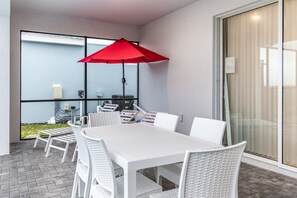 A modern outdoor dining area with a white table and chairs, a red umbrella, and a glass partition with a view of greenery. - Themed Rooms, Game Loft, & Private Pool Near Parks (Davenport)