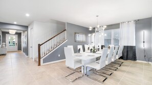 A modern dining area with a white table set for six, a staircase, and a view of the kitchen. - 9BR Lake View Frozen, Batman & More (Kissimmee)