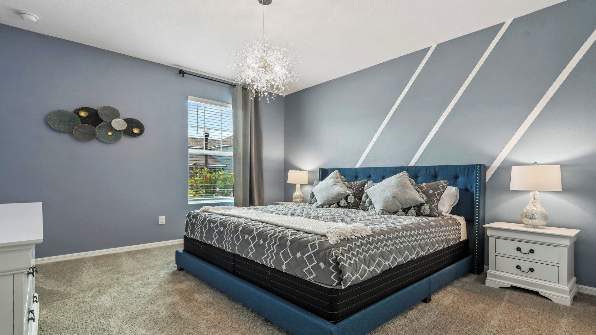 A bedroom with a large bed, two bedside tables, a chandelier, and a window with blinds.