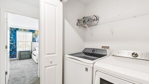 A laundry room with a front-loading washer and dryer, a drying rack with folded towels, and a door leading to a bedroom with a bed and a blue wall with a nature scene. - 4BR w Screened Pool & Resort Access Near Parks (Kissimmee)