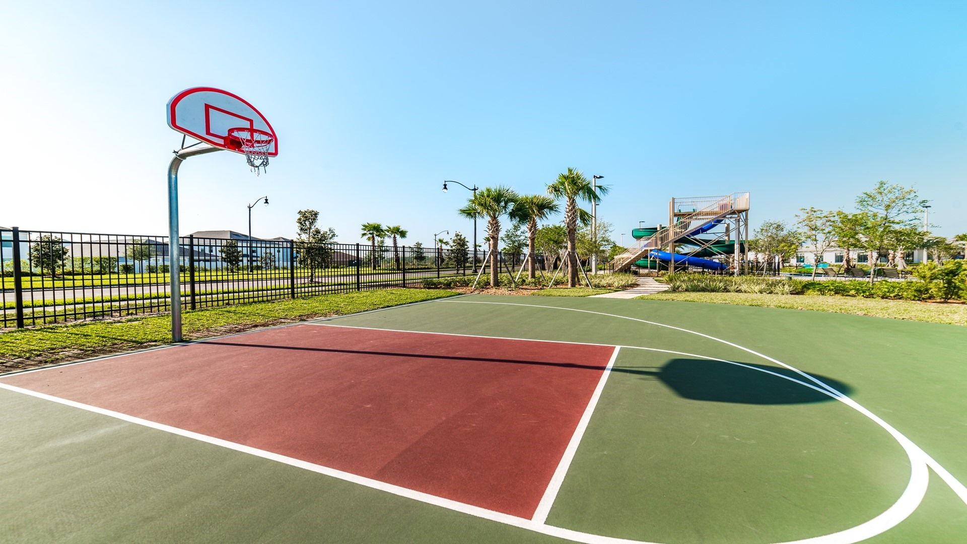 Ein Basketballplatz im Freien mit roter und grüner Spielfläche, umgeben von einem schwarzen Zaun, Palmen und einer Wasserrutsche im Hintergrund.