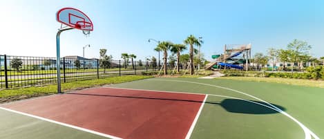 An outdoor basketball court with a red and green playing surface, surrounded by a black fence, palm trees, and a water slide structure in the background.