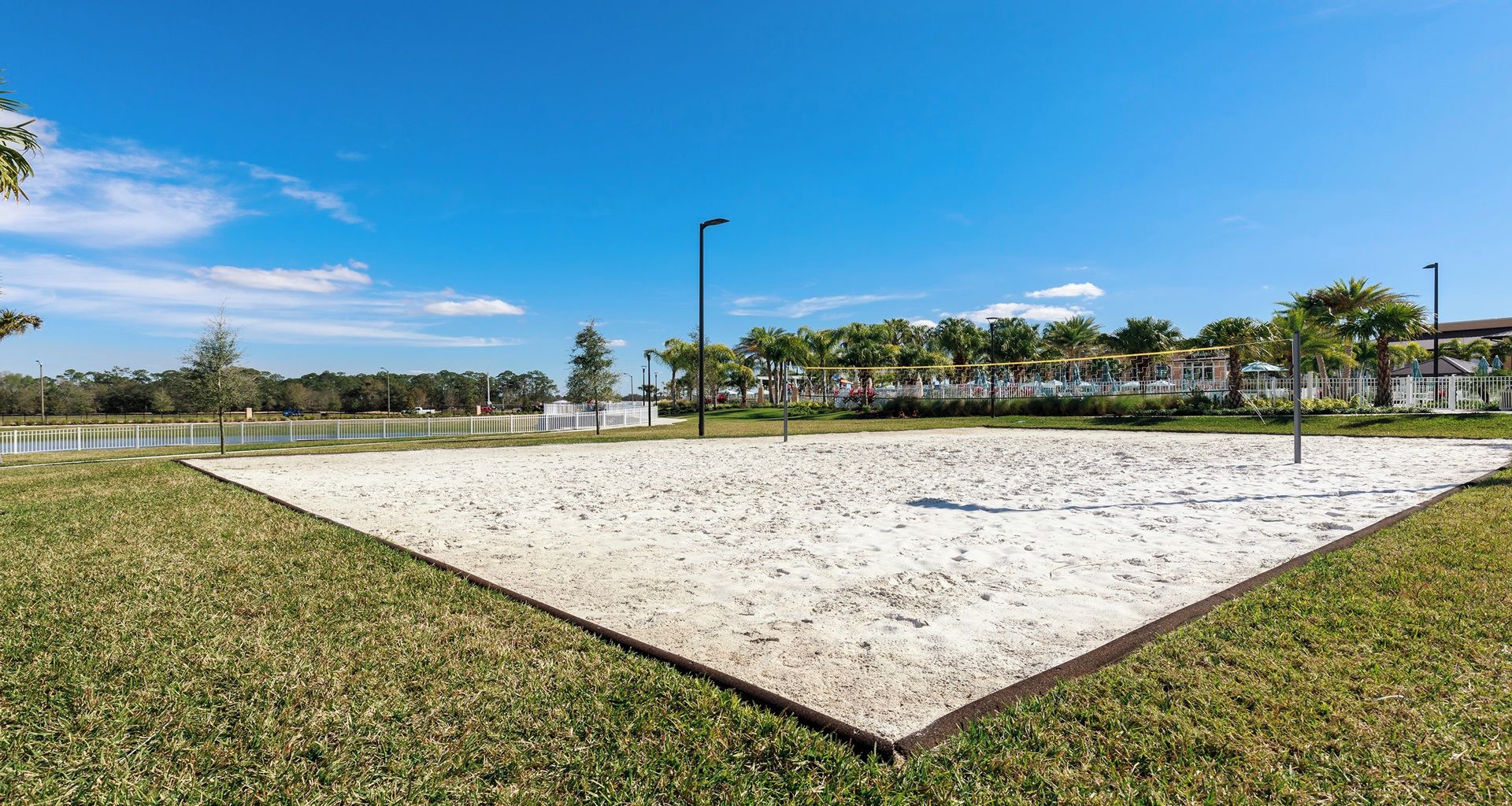 A sandy volleyball court with a surrounding fence, palm trees, and a clear blue sky.