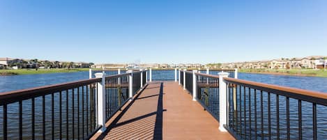 A wooden pier extending over water with railings on both sides, leading to a residential area with houses in the background.