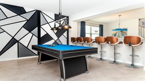 Een pooltafel met een blauwe viltkleed, omringd door barstoelen met houten zitvlakken en metalen poten.