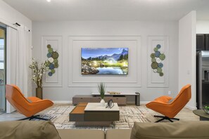 A modern living room with a large flat-screen TV mounted on the wall, two orange chairs, a coffee table, and decorative wall art. - Discounted 12br w Barbie Castle/dino Slide/games (Kissimmee)