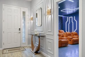 Un couloir avec une porte blanche, une console dont le plateau est en verre et un coin salon avec des fauteuils orange.