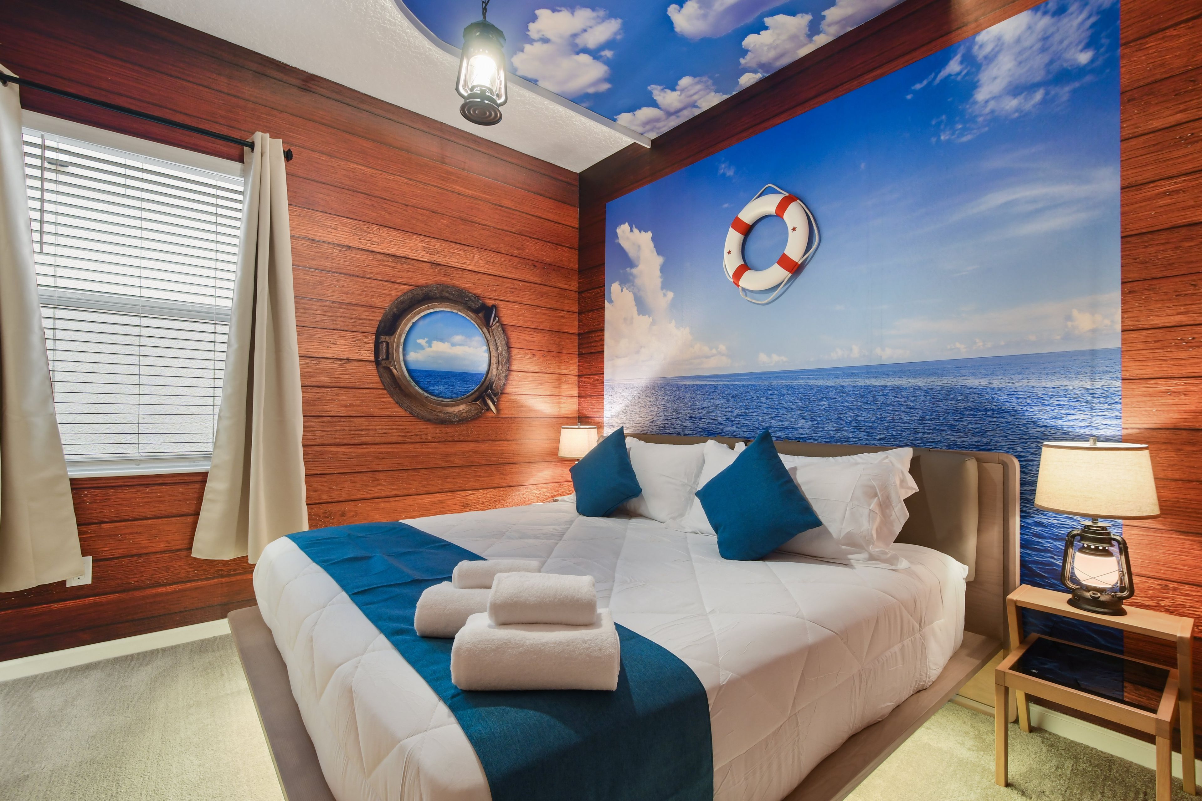 A bedroom with a bed, a nightstand, a lamp, a life preserver, a round mirror, and a window with curtains.