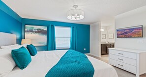 Ein Schlafzimmer mit Bett, Nachttisch, Fenster mit Vorhängen, einem Bild an der Wand und einem durchgegangenen Tür einsehbaren Badezimmer.