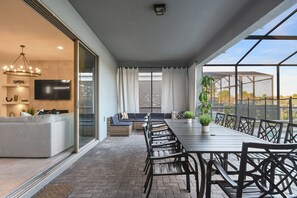 Un espace de repas extérieur moderne, avec une longue table et des chaises, un canapé et un téléviseur placé à gauche.