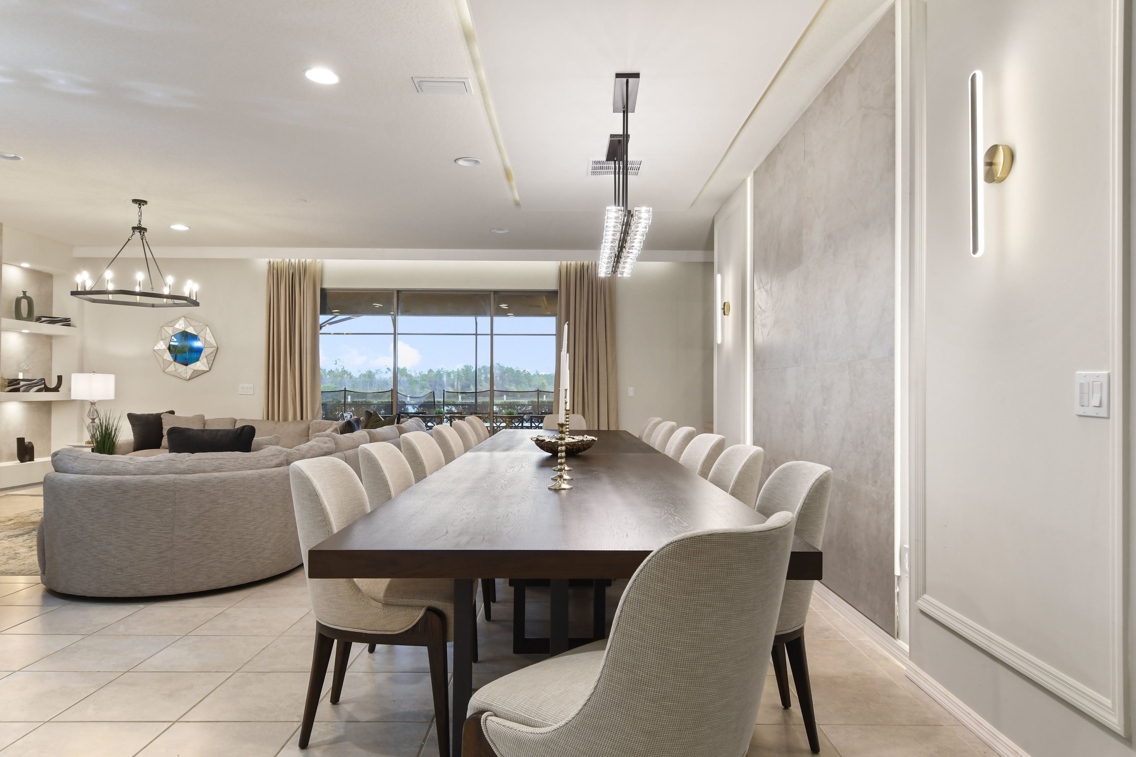 A modern dining area with a long table, multiple chairs, a chandelier, and a view through large windows.