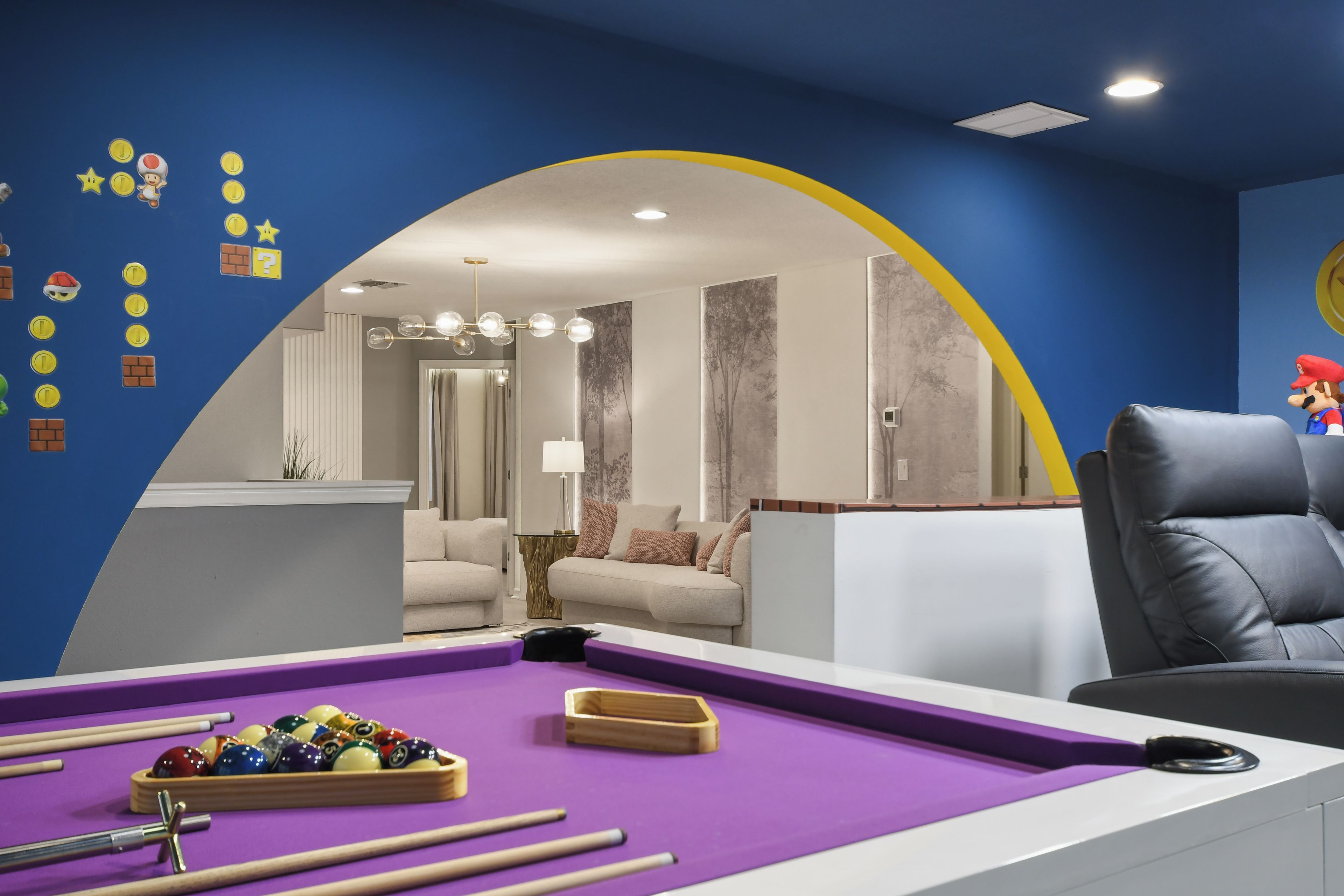 A pool table with balls and cues, a sofa, and a Mario figurine in a room with blue walls and a yellow arch.