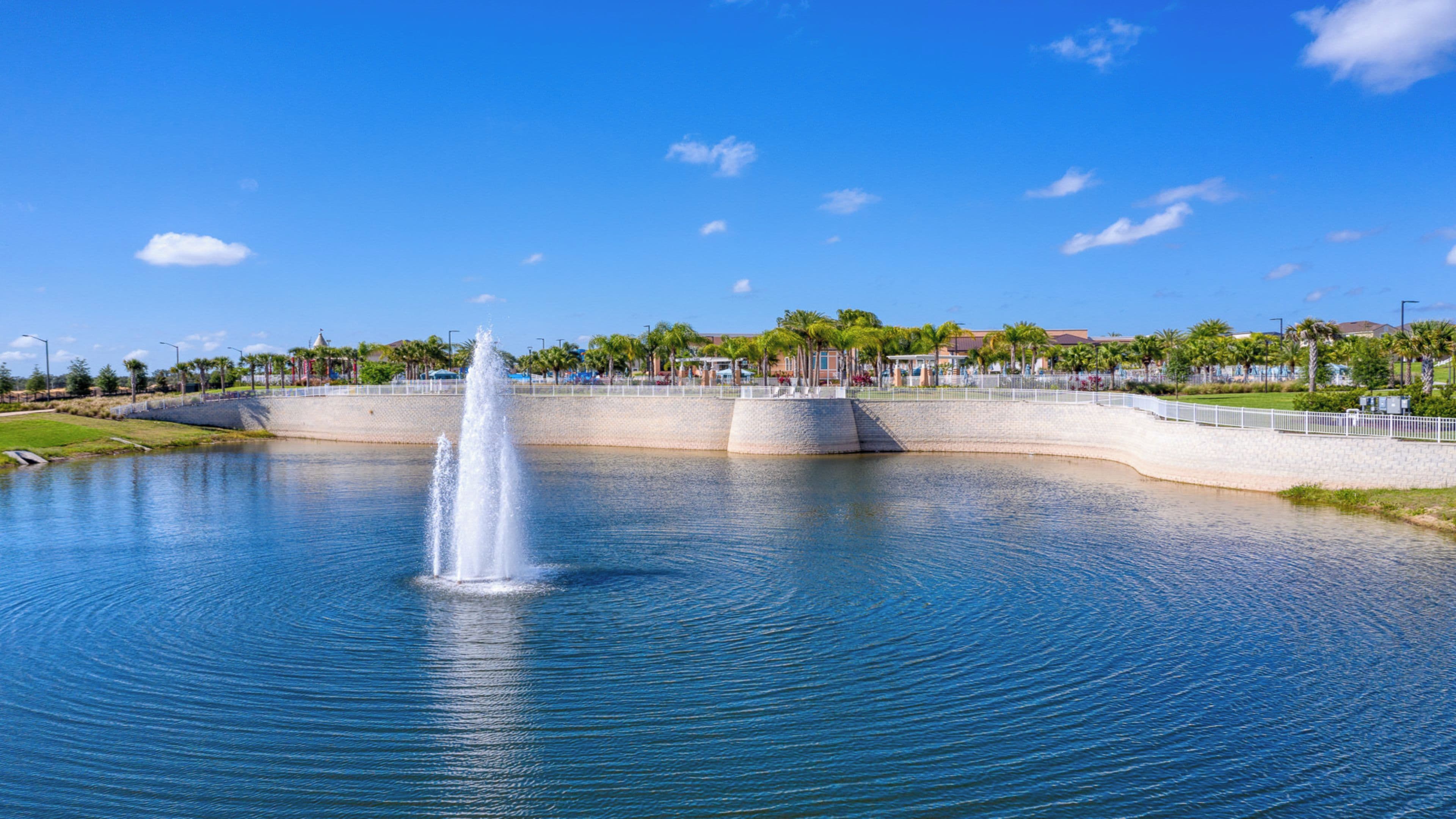 A large body of water with a central fountain, surrounded by a low stone wall and palm trees.