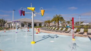 Ein Wasserpark mit verschiedenen Wasserspielen, darunter eine Wasserrutsche und Springbrunnen. Im Hintergrund sind Liegestühle und ein Gebäude zu sehen.