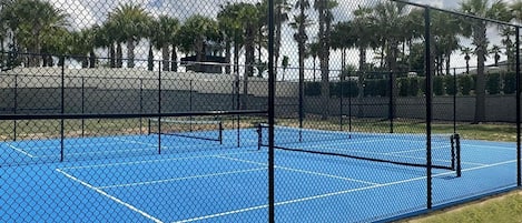 Cancha de tenis azul rodeada por una cerca de malla metálica, con palmeras al fondo.