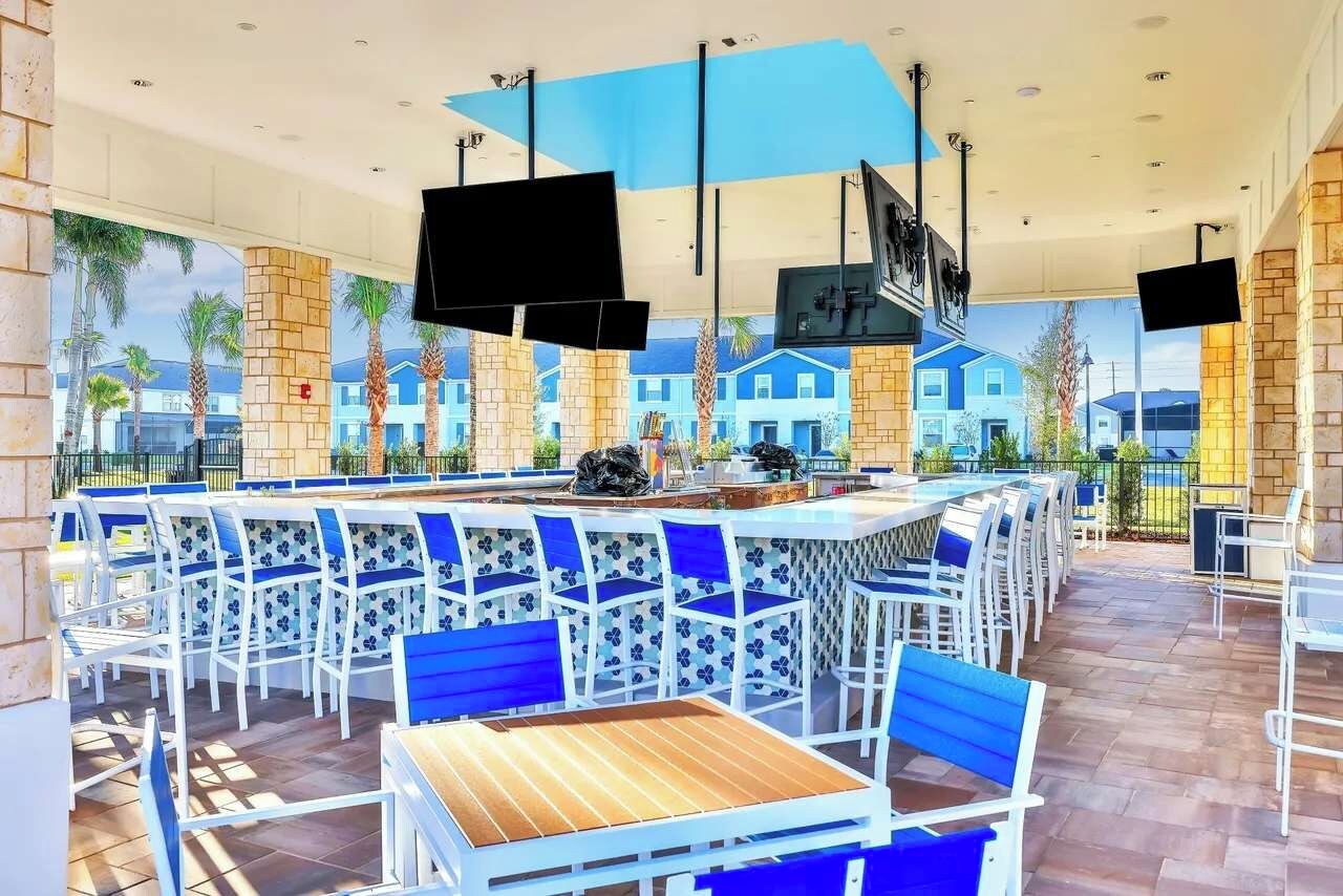 Un espace bar extérieur moderne, avec des chaises blanches et bleues, un comptoir central et de grands écrans fixés au plafond.