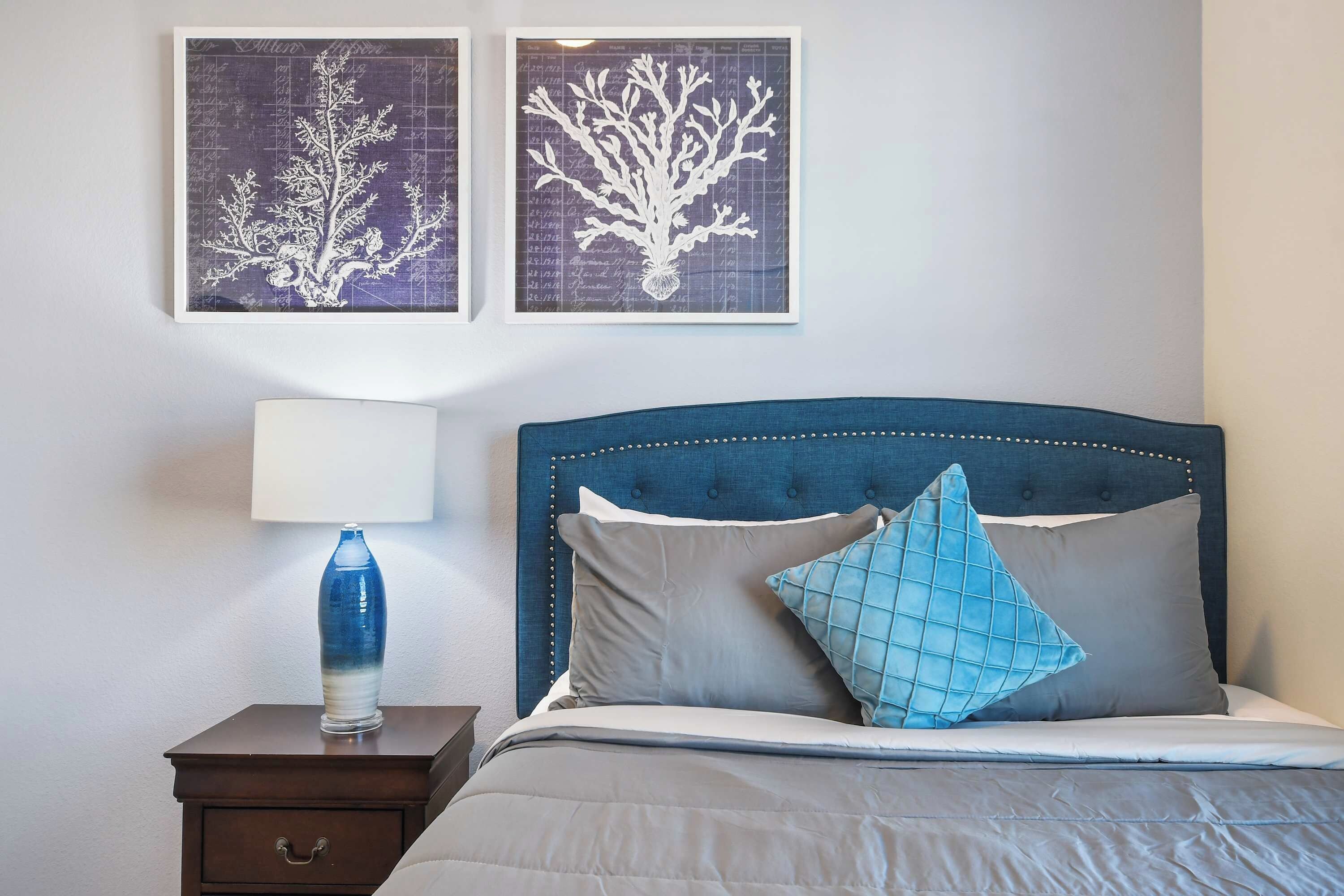 Una cama bien hecha con un cabecero azul, una lámpara de noche y dos impresiones enmarcadas de motivos botánicos en la pared.