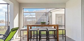 Eine sonnendurchflutete Terrasse mit einem Holztisch und vier Stühlen, ein grüner Sessel sowie eine Glastür mit Blick auf ein Haus und ein Gewässer.