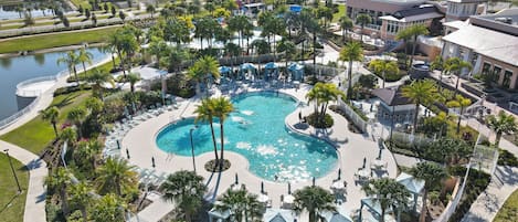 A large, circular swimming pool surrounded by palm trees and lounge chairs, with a view of nearby residential buildings.