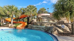 Une piscine avec un toboggan aquatique, des palmiers et une cascade artificielle en roches.