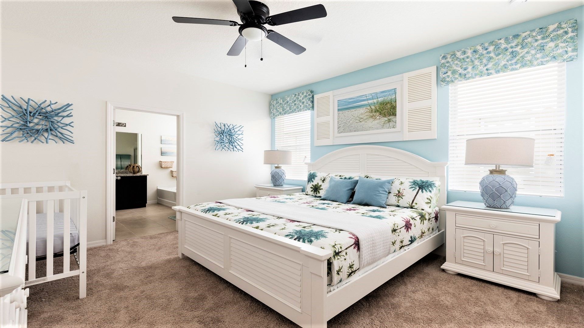 Quarto com cama, mesas de cabeceira, berço, ventilador de teto e arte decorativa na parede.