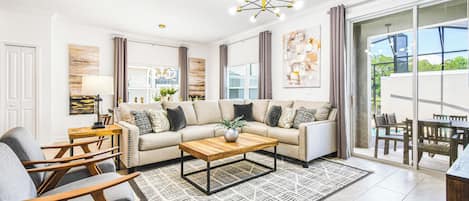 A modern living room with a sofa, coffee table, and chairs, a dining area with a table and chairs, and a large sliding glass door leading to an outdoor patio.