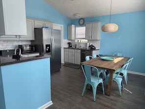 A modern kitchen with white cabinetry, a stainless steel refrigerator, and a wooden dining table with blue chairs. - Beautiful 3-bedroom house just 1 block from the beach in Panama City Beach (Panama City Beach)