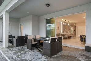 A modern outdoor dining area with wicker furniture and a glass table, leading to an interior dining space with a view of the outside. - Minion Themedprivate Pool Game Room & Green Views (Davenport)