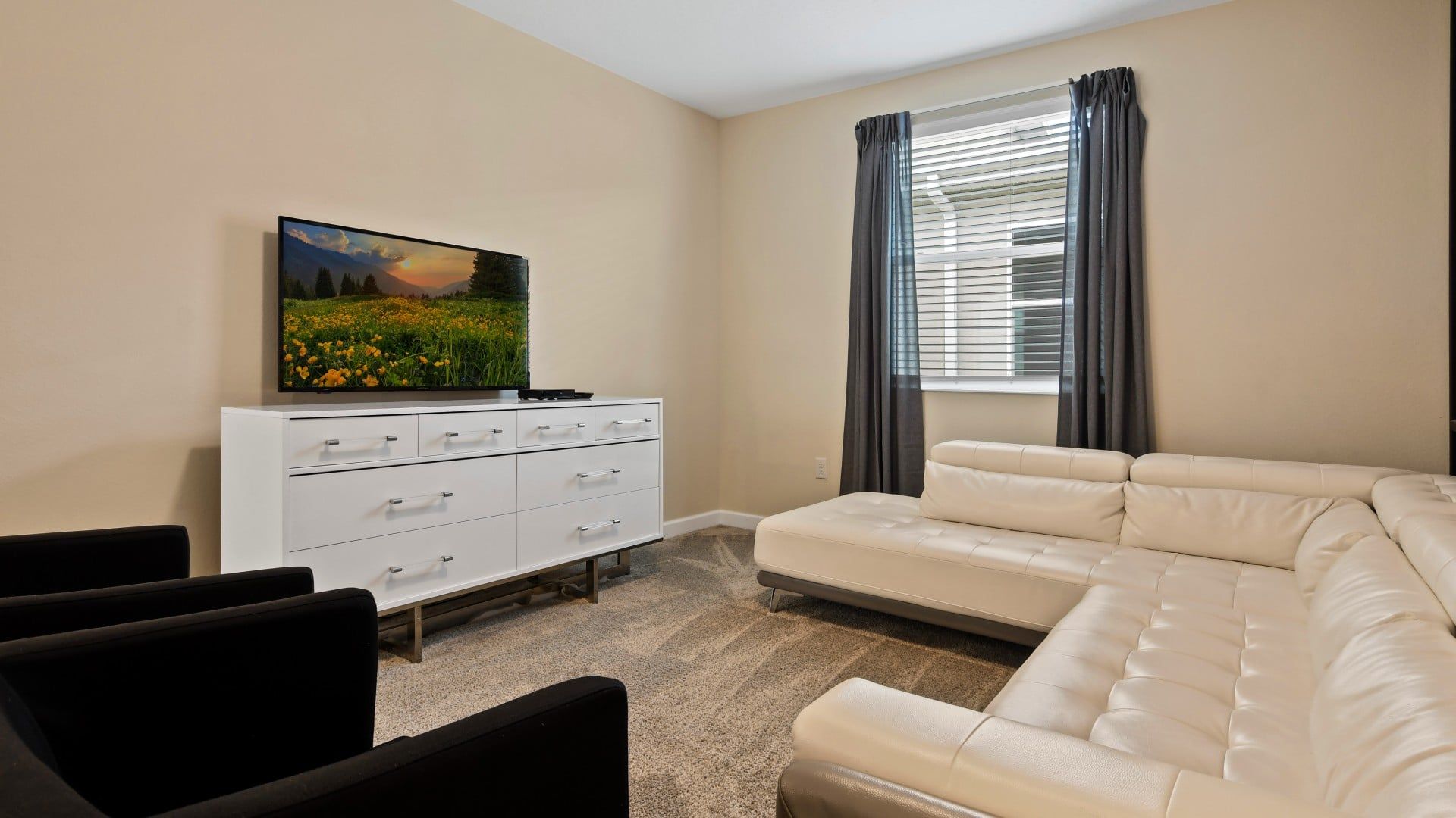 A living room with a white sofa, a flat-screen TV on a white cabinet, and a window with curtains.