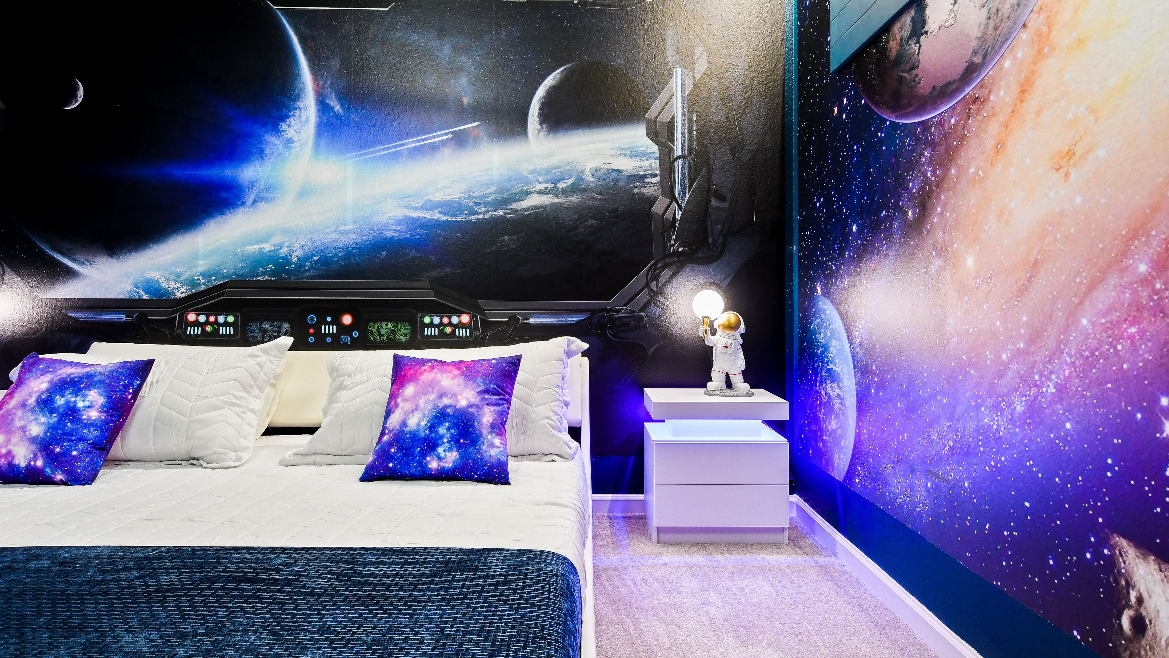 Une chambre dotée d’une grande fresque murale sur le thème de l’espace, d’un lit avec une literie blanche et des oreillers à motifs galactiques, d’une table de chevet avec une petite lampe, et offrant une vue sur la Terre depuis l’espace.
