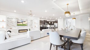 A modern living room with a fireplace, a dining area with a large table and chairs, and a kitchen with dark cabinets and a stainless steel refrigerator. - Mermaid & Snow White-themed Escape Near Disney (Davenport)