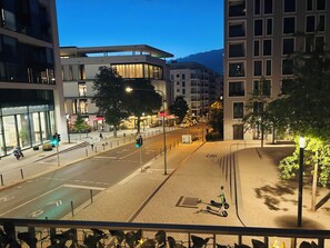 A city street at dusk with modern buildings, a pedestrian crossing, and a scooter parked on the sidewalk.