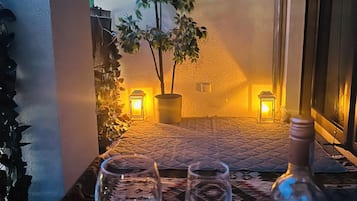 Une table avec deux verres à vin et une bouteille de vin, une plante en pot et deux lampes fixées au mur, dans une pièce dont le mur est texturé et qui possède une fenêtre.