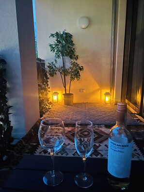 Une table avec deux verres à vin et une bouteille de vin, une plante en pot et deux lampes fixées au mur, dans une pièce dont le mur est texturé et qui possède une fenêtre.