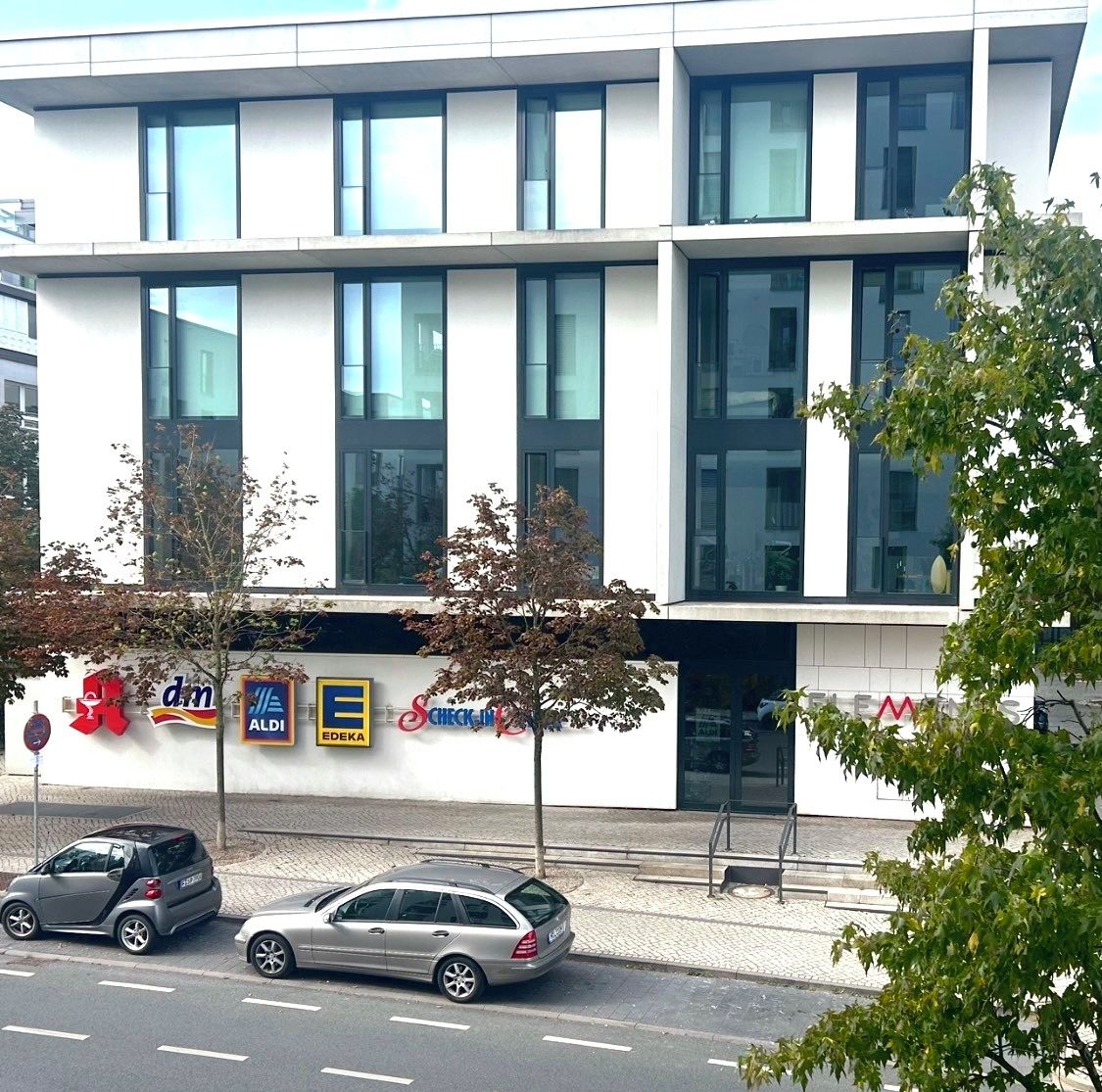 Ein modernes Gebäude mit großen Fenstern und einer weißen Fassade. Es gibt mehrere Geschäfte mit sichtbaren Logos, darunter ein Autohaus und eine Check-in-Service.