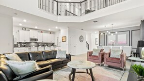 A spacious living room with a black leather sofa, a round coffee table, and two pink armchairs with fluffy pillows. A kitchen with white cabinets and a microwave is visible in the background. - Hogwarts to Arendelle: Themed Magic Near Disney (Kissimmee)