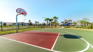 Ein Basketballplatz im Freien mit roter und grüner Spielfläche, umgeben von einem Zaun, Palmen und einer Wasserrutsche im Hintergrund.