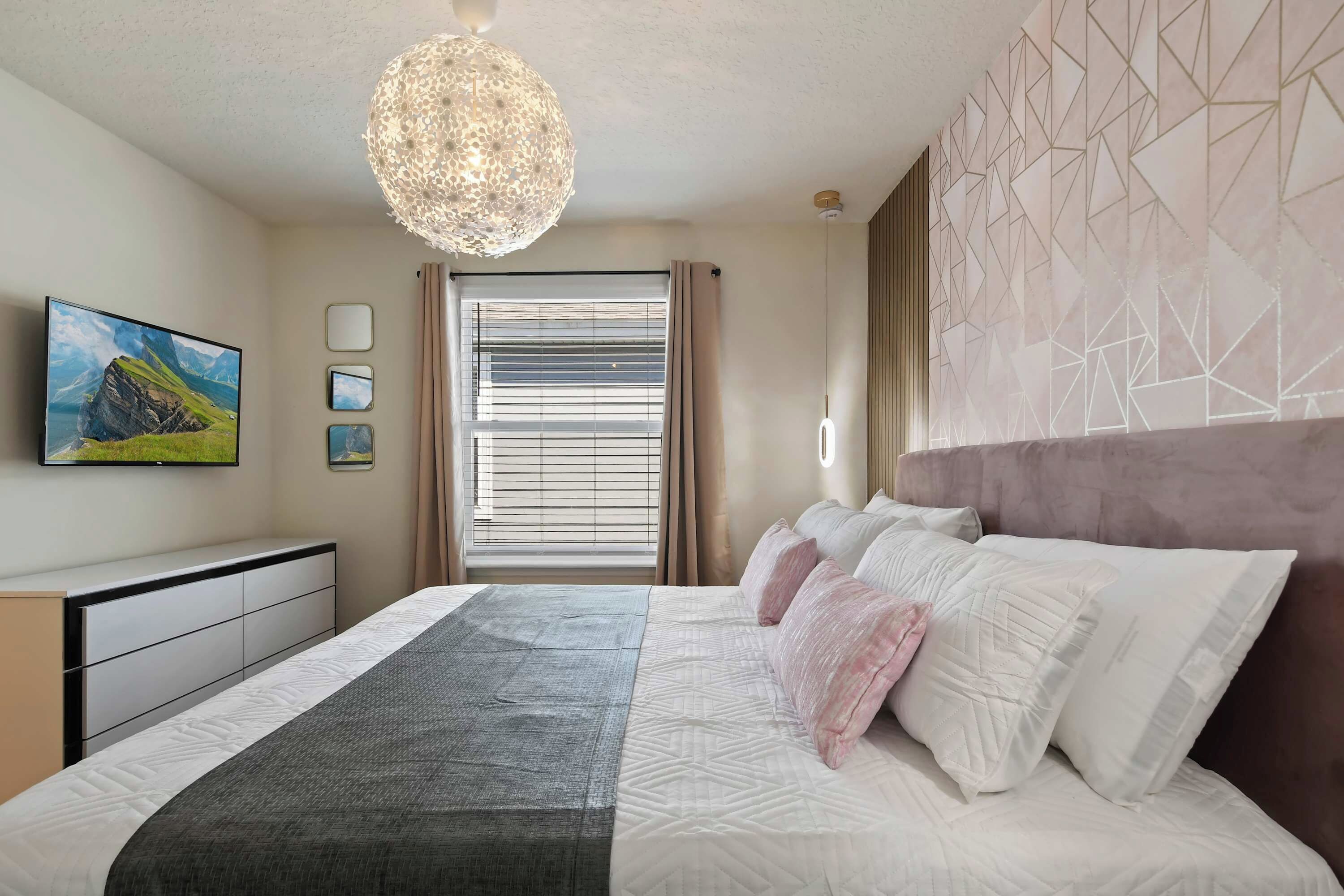 A bedroom with a bed, a large window with blinds, a wall-mounted TV, and a geometric-patterned wall.