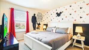 Una camera d'albergo con un letto, una TV, una finestra con tende e una parete decorata con l'immagine di Topolino, oltre a una frase motivazionale.