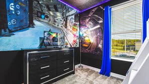 A room with a large TV displaying a movie, a desk with drawers, a window with blue curtains, and a wall with a space-themed mural. - Disney! Themed Rooms, Private Pool, Resort Access! (Kissimmee)