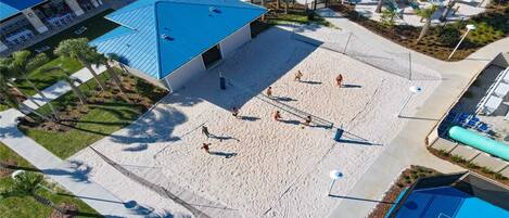 Un terrain de beach-volley avec des joueurs, une piscine et un bâtiment avec un toit bleu.