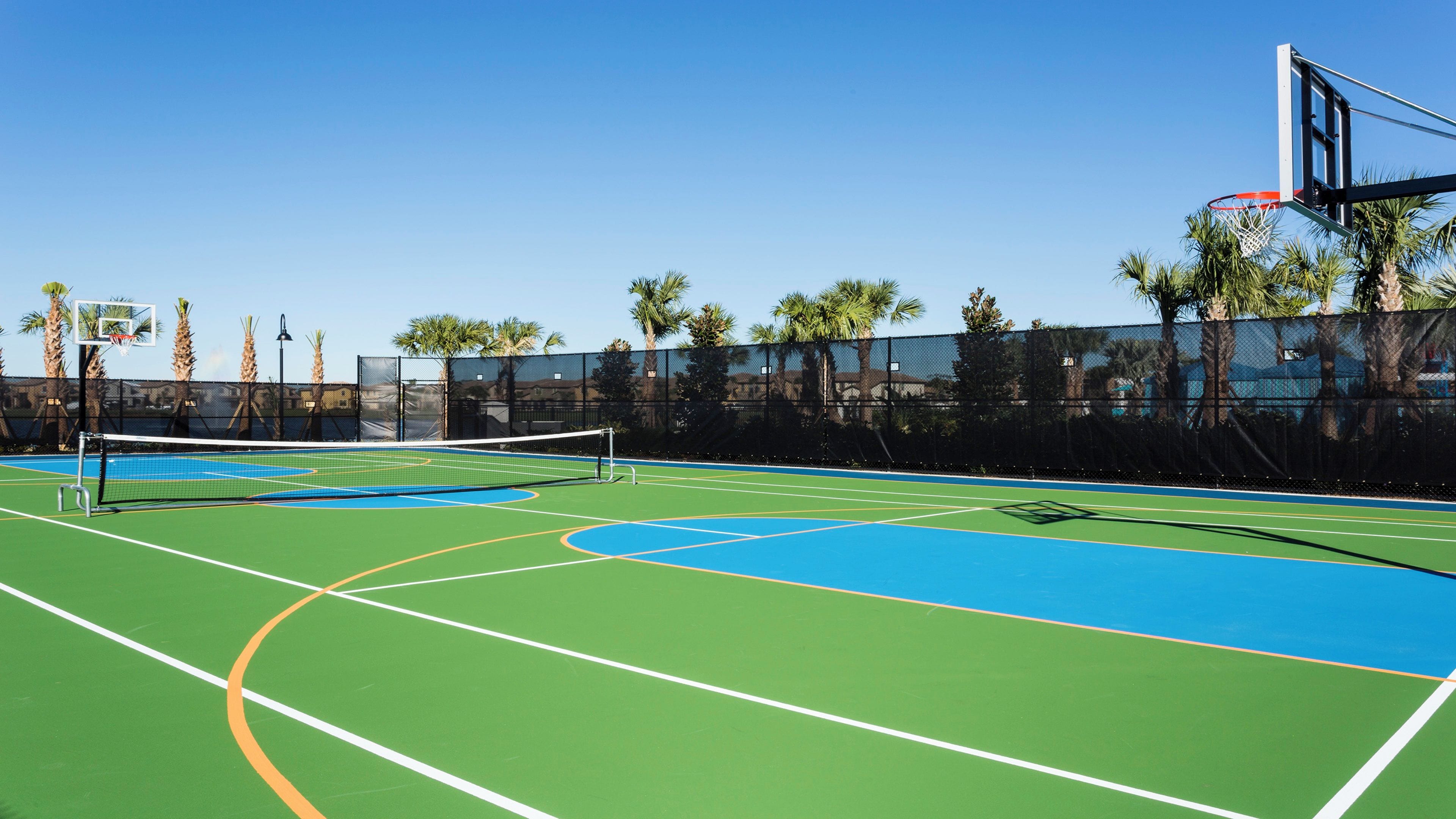 An outdoor tennis court with a net, surrounded by a fence and palm trees.